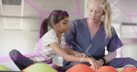 Helping therapist guiding girl's arm in bright rehab room with navy scrubs smartwatch foam cushions. Pediatric, clinician, movement, rehabilitation, therapy, windows, naturalの写真素材
