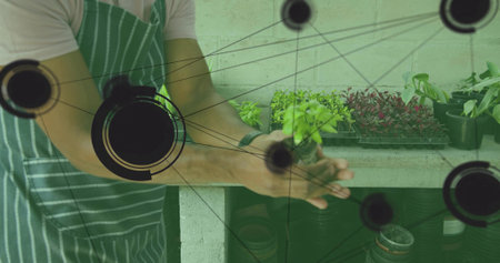Gardener wearing apron inspecting potted seedling at greenhouse bench, with trays and data overlay....