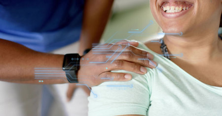 Smiling seated woman wearing mint top, receiving shoulder touch in clinic with black smartwatch HUD. Caregiving, support, healthcare, technology, overlay, connection, comfortの写真素材