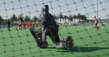 Kneeling field hockey goalkeeper wearing helmet and leg guards defending net on turf, with cones. Sports, competition, athletic, training, teamwork, determination, outdoorsの写真素材