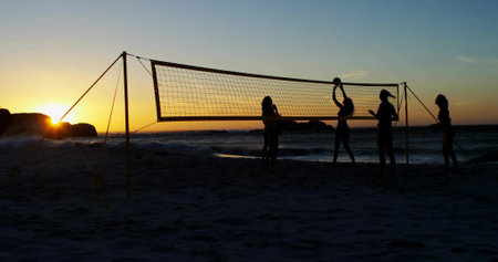 Playing five women wearing swimwear and cap spiking volleyball on beach at sunset, with net. Silhouettes, shoreline, dusk, waves, friends, leisure, horizonの写真素材