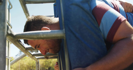 Placing man in striped polo slipping through metal frame outdoors, with blue pad and gripping hand. Rails, rigging, sunlit, shadows, nature, sky, focusの写真素材