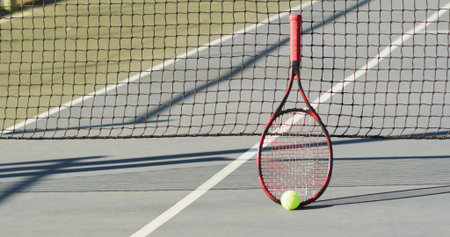 Leaning red tennis racket holding yellow tennis ball against net on open court, with boundary lines. Athletic, sport, competition, outdoor, vibrant, leisure, trainingの写真素材