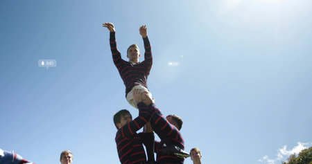 Lifting rugby teammate during lineout wearing striped jerseys, beige shorts on grass under blue sky....