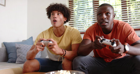 Playing two men on sofa gripping white controllers in living room, mustard and orange T-shirts. Gaming, friends, casual, home, daylight, cozy, competitionの写真素材