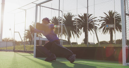 Diving teenage female goalkeeper in full gear lunging on turf with goal frame, helmet, sun flare. Keeper, fieldhockey, artificialgrass, backlit, goldenhour, palmtrees, chainlinkの写真素材