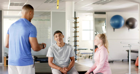 Standing trainer in blue shirt holding timer and instructing seated clients on treatment table, TRX. Rehab, therapy, gym, equipment, guidance, assessment, brightの写真素材