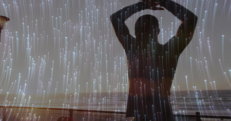Stretching man raising arms overhead on boardwalk by metal railing at dusk, with digital light. Meditation, wellness, contemplation, serenity, silhouette, relaxation, modernの写真素材