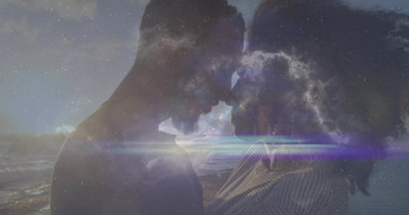 Touching foreheads couple standing by waves at dusk, with clouds, horizon, star-specks, light flare. Romantic, connection, serenity, silhouette, dusk, tranquility, artistryの写真素材