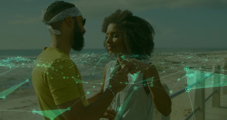 Standing couple facing each other on sandy beach with sunglasses, bandana, metal railing overlay. Romance, outdoor, scenic, leisure, connection, harmony, vibrantの写真素材