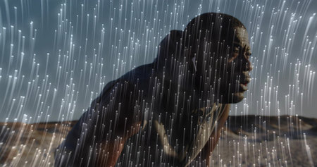 Crouching athletic man in dark sleeveless top leaning forward in desert dunes, with luminous rain....