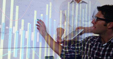 Team in business attire analyzing bar chart on glass board in conference room, with marker, tablet....