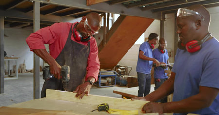 Drilling carpenter in red shirt securing plank at workshop bench, with cordless drill, copy space. Craftsmanship, teamwork, industrial, manufacturing, hands-on, construction, texturedの写真素材