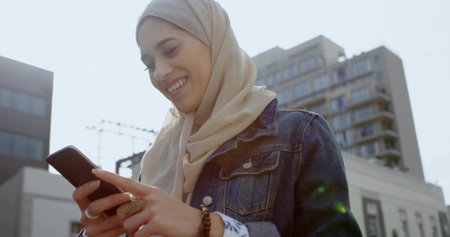 Smiling woman wearing scarf and denim jacket checking smartphone in city plaza with ring, bracelet....
