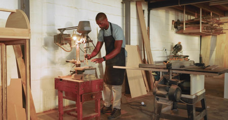 Mature African American carpenter feeding lumber into drill press in woodshop, with table saw. Craftsman, woodworking, industrial, rustic, craftsmanship, professional, toolsの写真素材