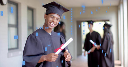 Smiling male teen in black gown, mortarboard cap holding diploma in school corridor, with windows....