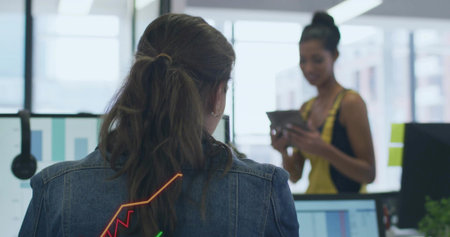 Denim-jacketed woman analyzing charts on monitors in open office, with colleague glancing at tablet. Collaboration, analytics, technology, workspace, productivity, modern, professionalの写真素材