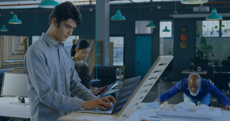 Typing man wearing gray shirt at high table in office, with laptop, architectural model, copy space. Industrial, collaboration, workspace, modern, design, teamwork, environmentの写真素材