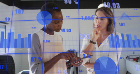 Examining tablet computer, two business women analyzing data in modern office with floating charts. Collaboration, analytics, business, modernity, data visualization, professionalism, teamworkの写真素材