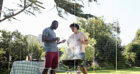 Grilling two men tending charcoal grill in yard holding sauce bottle and tongs near checked table. Barbecue, outdoor, friends, vibrant, picnic, casual, leisureの写真素材