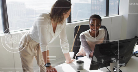 Collaborating two mid adult women wearing blouses leaning over PC at office desk, with coffee mug. Collaboration, teamwork, modern, professional, workspace, cityscape, businessの写真素材