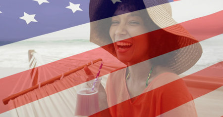 Relaxing woman wearing straw hat holding red drink in hammock on beach, with American flag overlay. Tropical, leisure, summer, tranquil, vibrant, outdoor, wellnessの写真素材