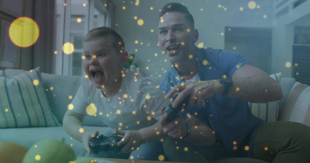 Cheering father and son playing console game on sofa in living room, with controllers, green apples. Family, bonding, entertainment, leisure, cozy, modern, happinessの写真素材