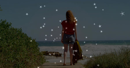 Standing woman holding jacket over arm facing ocean on wooden boardwalk, green shrubs, sandy beach. Coastal, serenity, leisure, landscape, outdoor, nature, relaxingの写真素材
