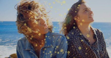 Gazing two women wearing denim jacket, patterned button-down shirt standing on rocky shore by ocean. Adventure, friendship, outdoor, nature, vibrant, carefree, explorationの写真素材