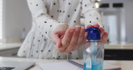 Pumping woman in polka-dot blouse dispensing sanitizer from bottle in kitchen, notebook, laptop. Hygiene, cleanliness, health, protection, wellness, minimalism, modernの写真素材