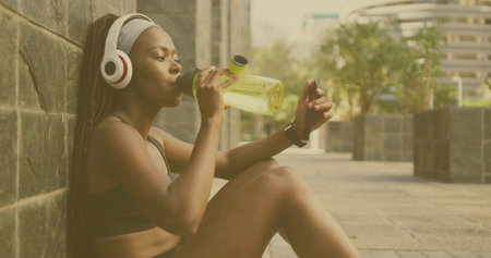 Sitting fitness fan in sportswear drinking from water bottle on plaza, with headphones, copy space. Athletic, active, urban, wellness, lifestyle, modern, vibrantの写真素材
