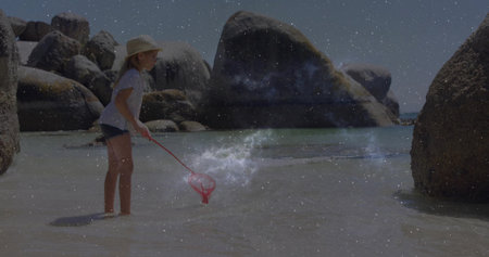 Scooping water child wearing straw hat, shorts at rocky beach cove with red net, copy space. Youth, exploration, leisure, nature, adventure, tranquility, playfulの写真素材