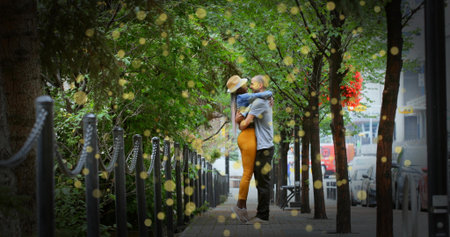 Couple embracing and lifting in sidewalk, with mustard outfit and hat, bollards, cars, golden bokeh. Romance, affection, urban landscape, dreamy, joy, connection, fashionの写真素材