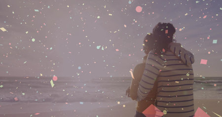 Embracing couple in tank top, striped hoodie watching sunset at beach, confetti overlay, copy space. Romance, celebration, serenity, affection, harmony, joy, leisureの写真素材