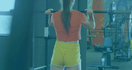 Woman in orange top and yellow shorts performing squat in gym weight area, with barbell. Fitness,...