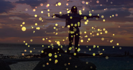 Standing female silhouette spreading arms on rocky shoreline at sunset, with golden bokeh lights. Serenity, inspiration, tranquility, wellness, sunset, ethereal, natureの写真素材