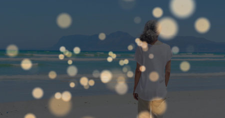 Standing senior woman gazing toward horizon at sandy beach, with golden bokeh lights, copy space. Serenity, contemplation, leisure, elegance, tranquility, nature, minimalistの写真素材