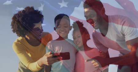 Standing family of four wearing T-shirts sharing smartphone outdoors, with American flag overlay. Patriotism, unity, celebration, diversity, technology, happiness, communityの写真素材