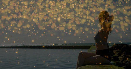 Sitting athlete wearing sportswear on rocky ledge by water at sunrise with golden orbs, copy space. Fitness, serenity, nature, dawn, tranquility, outdoor, inspirationの写真素材