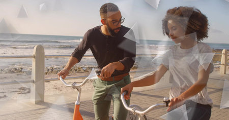 Walking couple pushing orange bicycles along seaside promenade by sandy beach, with metal railing....