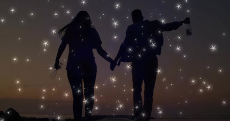 Walking backlit couple holding hands along shore at dusk, with glass bottle, sandals and sparkles....