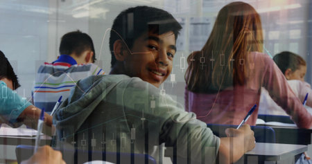 Smiling teenage boy in gray hoodie holding pen above paper at classroom desks, with notebooks. Education, students, classroom, learning, youthful, academic, teamworkの写真素材