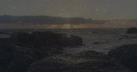 Sun sinking over horizon, casting glow on ocean waves crashing on rocky seashore under cloud layer....