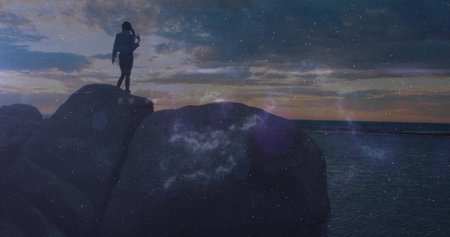 Standing silhouette shading eyes gazing horizon atop boulders beside calm water at dusk, copy space. Serene, contemplation, adventure, natural, tranquility, mountain, explorationの写真素材
