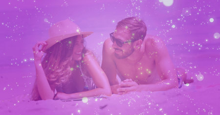 Smiling couple lying on sandy beach near shoreline, wearing swimwear with straw hat and sunglasses....