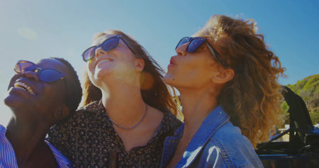 Leaning trio of friends in sunglasses tilting heads back in convertible by seaside road, sun flare. Adventure, camaraderie, summer, leisure, scenic, bright, outdoorの写真素材