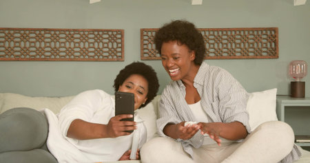 Holding black smartphone, two mid-adult women leaning, laughing on sofa at home with wooden panels....