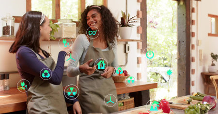 Smiling two women in gray aprons holding phone, gesturing at kitchen counter with produce and icons....