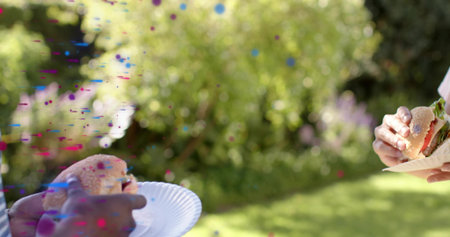 Two men wearing short sleeves holding burgers with lettuce on paper plates in sunny yard, confetti. Outdoor, backyard, picnic, friends, sesame, garden, vibrantの写真素材