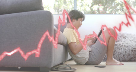 Sitting Asian man wearing pale yellow tee, holding tablet on hardwood floor, red graph overlay. Home, device, sofa, rug, naturallight, barefoot, casualの写真素材